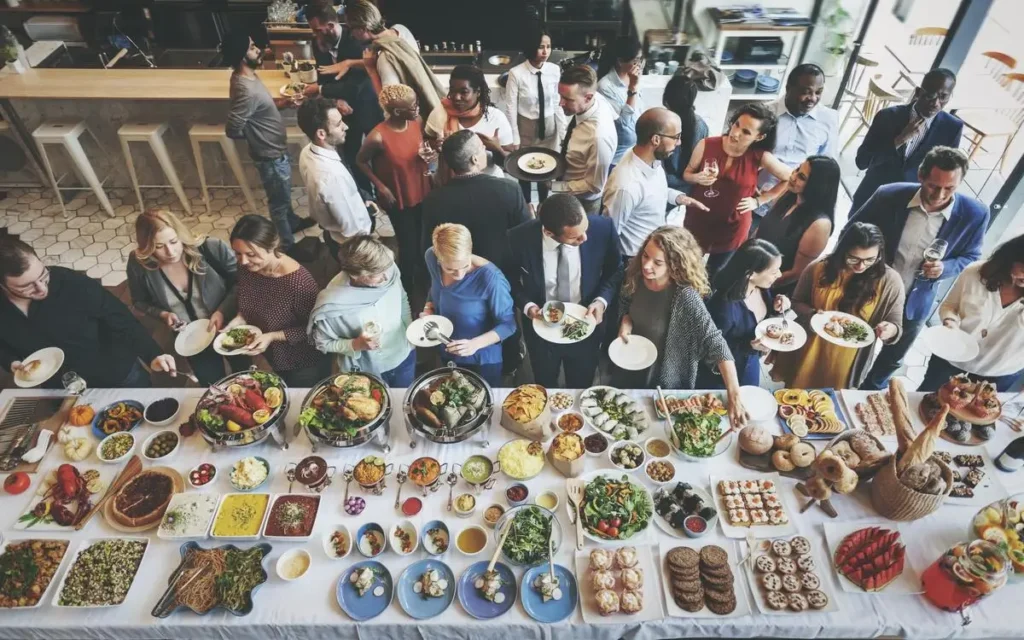 A bustling group of people serving themselves from a Mediterranean buffet spread at a large indoor event.