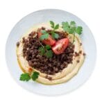 Hummus With Beef topped with seasoned halal ground beef, tomato wedges, parsley and olive oil on a white plate