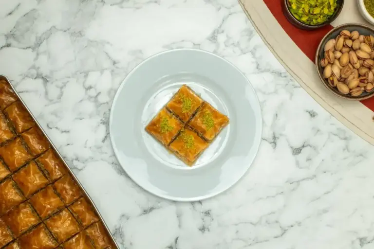 Pistachio baklava cut into diamonds, topped with ground pistachios, on a white plate beside a full tray and bowls of pistachios.